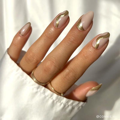 A hand with short, almond-shaped nails flaunts the glamnetic Material Girl set: a nude base with abstract gold foil swirls. The look is paired with two delicate gold rings and a white long-sleeved shirt for added chic style.